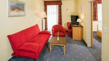 A hotel room with a red sofa, a small table with a vase, a wooden side table with a lamp, a television, and a bathroom visible through an open door.