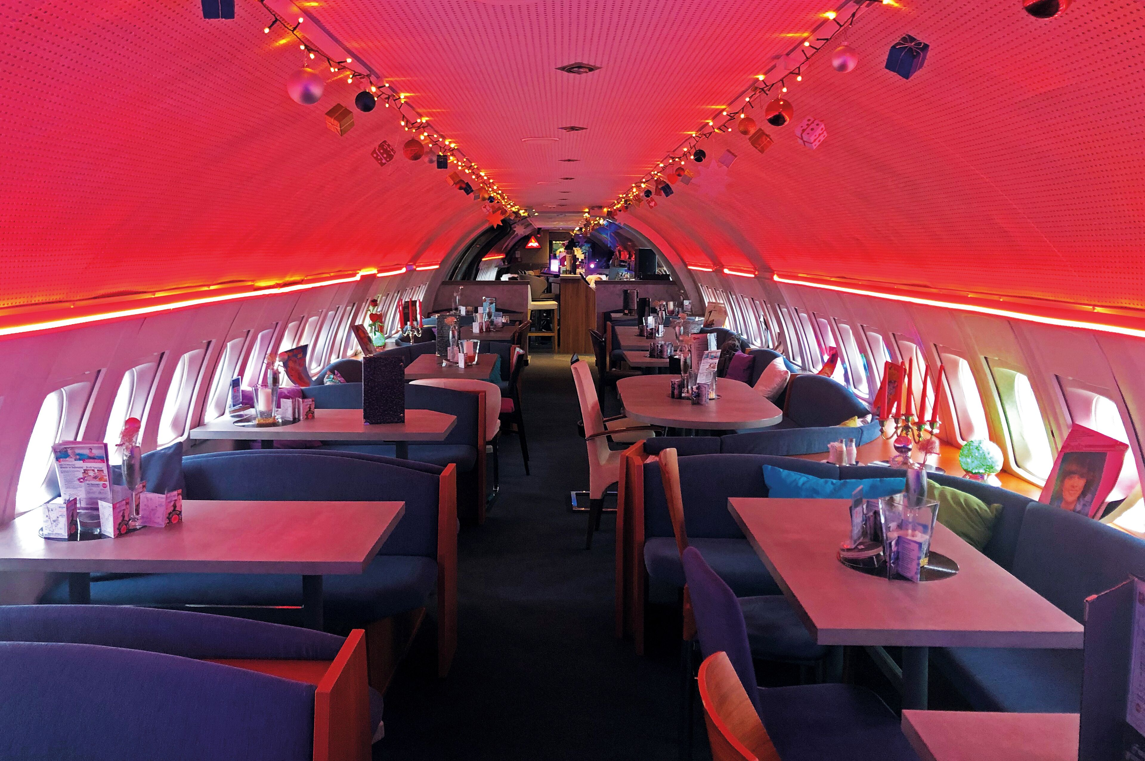 Interior de avión con mesas y asientos dispuestos en un círculo, iluminación en el techo y vistas del cielo a través de las ventanas.