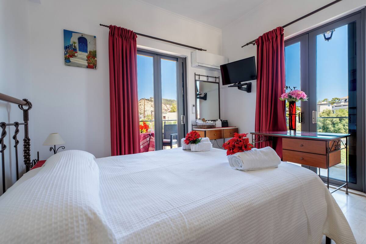 A hotel room with a large bed, a desk, a TV, and a balcony with red curtains.