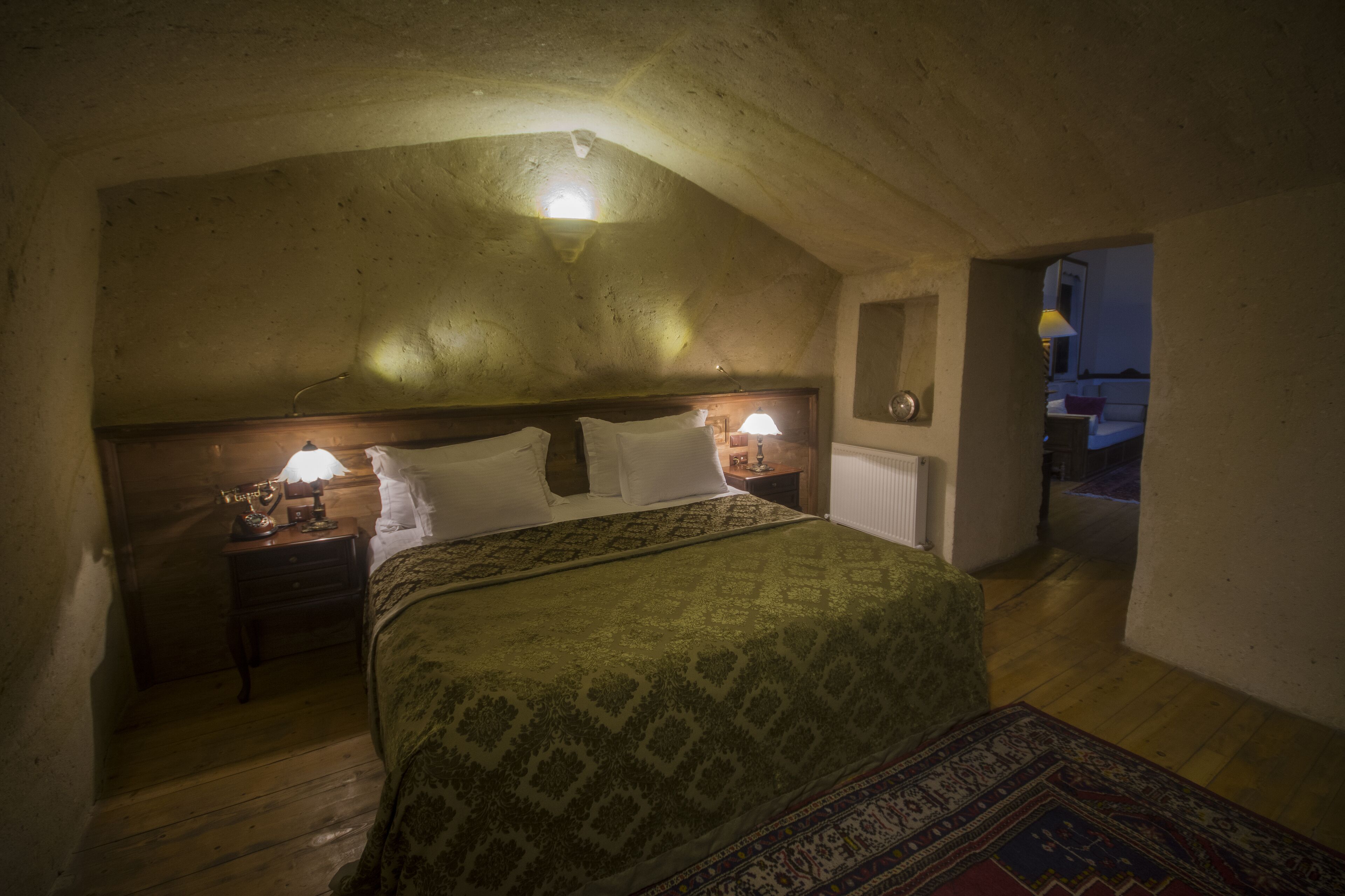 A bedroom with a wooden headboard, a bed with a patterned bedspread, two bedside lamps, and a nightstand with a lamp.