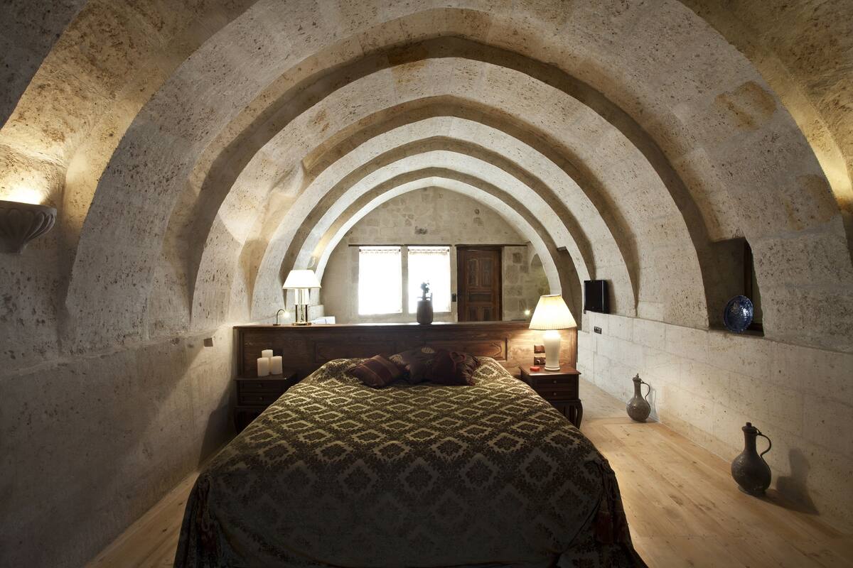 Un dormitorio con techo abovedado de piedra, una cama con colcha estampada, lámparas de noche y una mesita de madera.