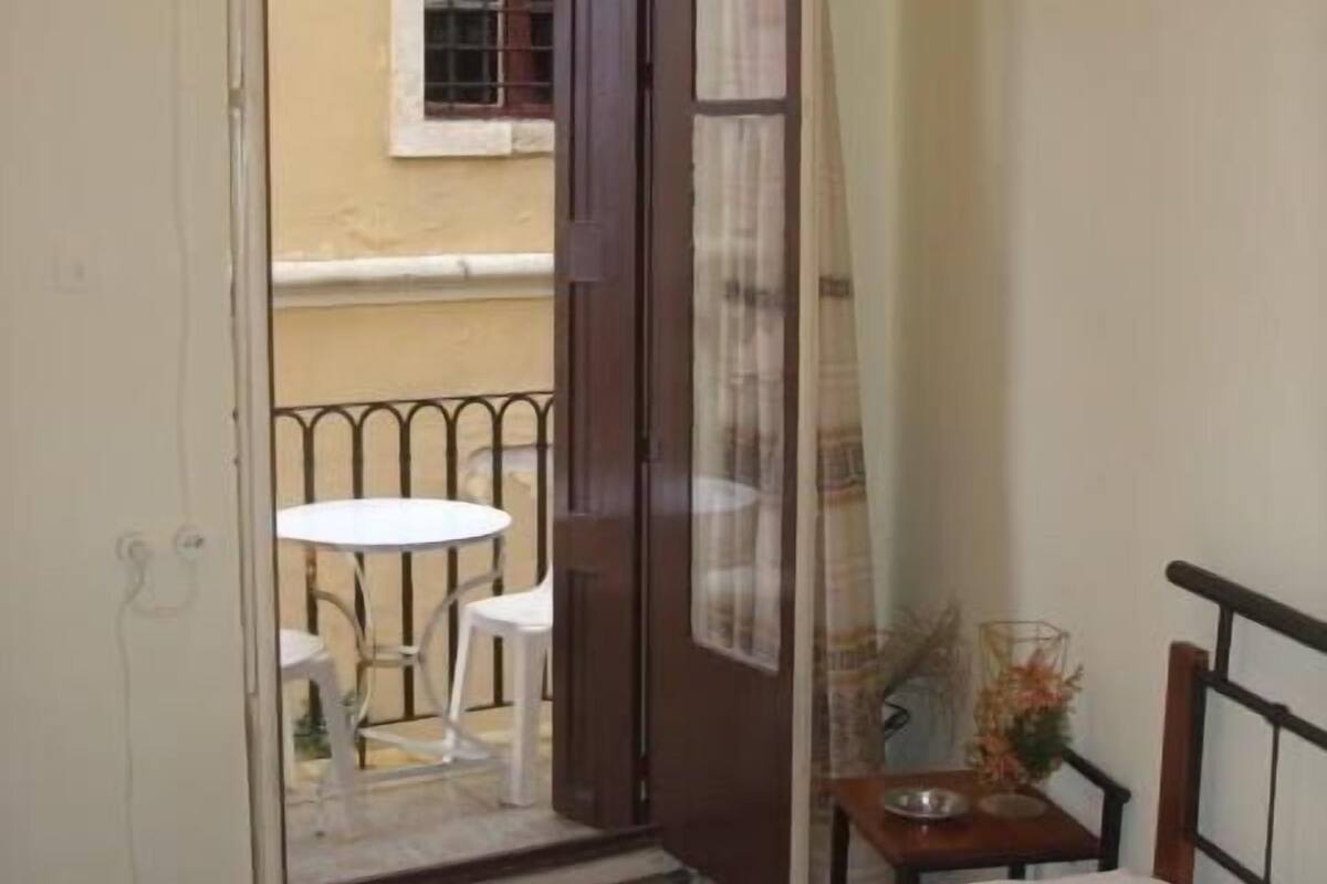 A bedroom with a bed, a small table with a vase of flowers, and a view of an outdoor balcony with a table and chairs.