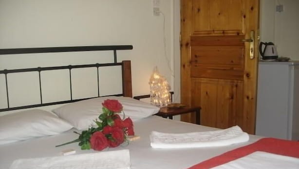 Una habitación de hotel con cama, puerta de madera, mesita de noche con lámpara y kitchenette.