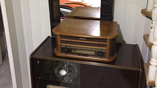 Une vieille radio et un vieux téléviseur sont posés sur un meuble en bois dans une pièce où se trouvent un lit et une porte en bois.