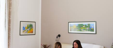 Une chambre à coucher avec un lit, deux personnes qui lisent, une couverture rouge et deux tableaux encadrés accrochés au mur.