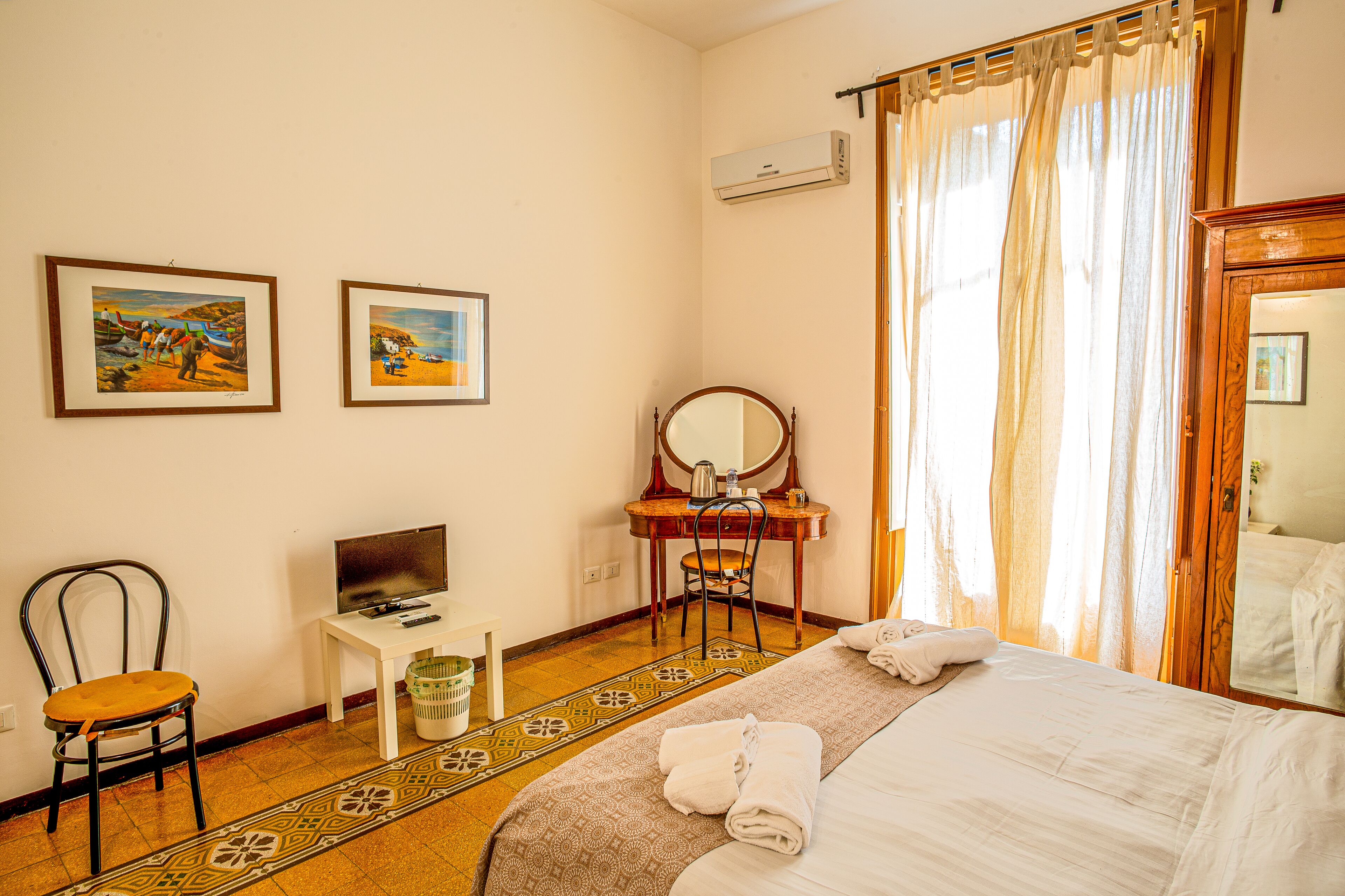 A hotel room with a bed, a desk with a computer, a chair, a mirror, and two framed pictures on the wall.