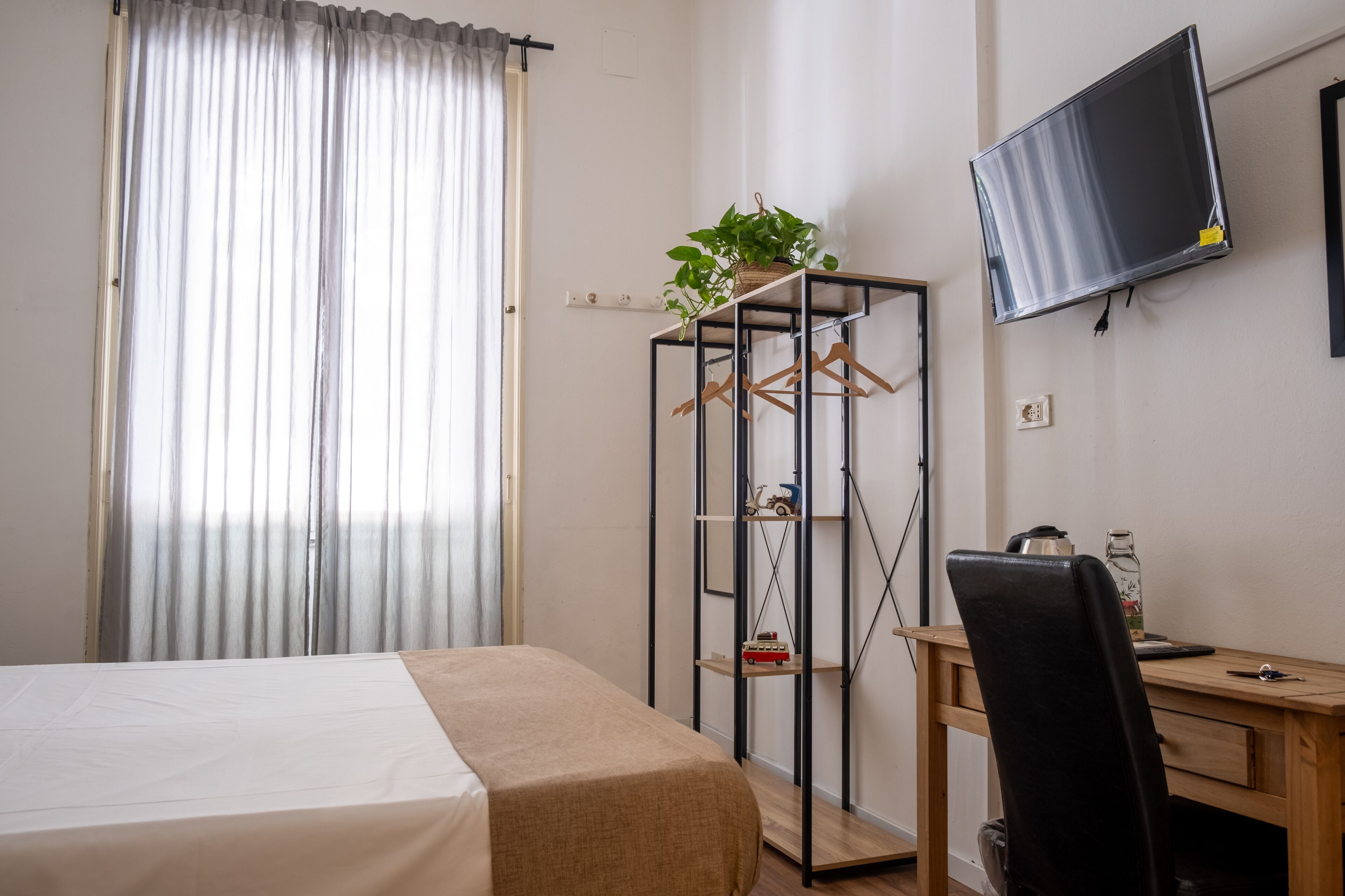 Une chambre à coucher avec un lit, un bureau avec une chaise, un téléviseur fixé au mur et une plante posée sur une étagère.