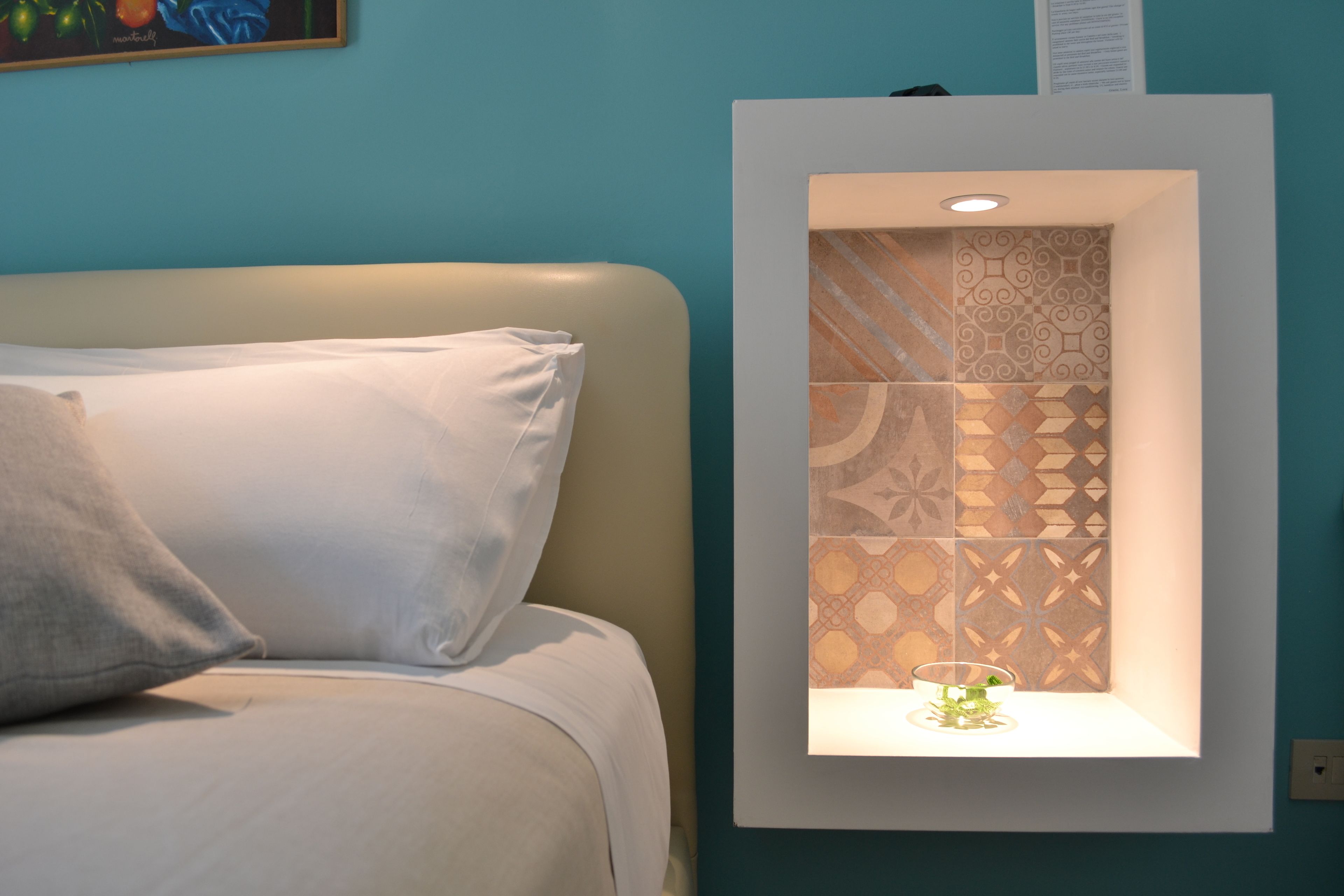 A bed with white bedding and pillows, a wall-mounted shelf with patterned tiles, and a small plant on the shelf.