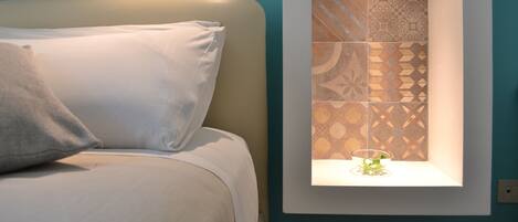 A bed with white bedding and pillows, a wall-mounted shelf with patterned tiles, and a small plant on the shelf.