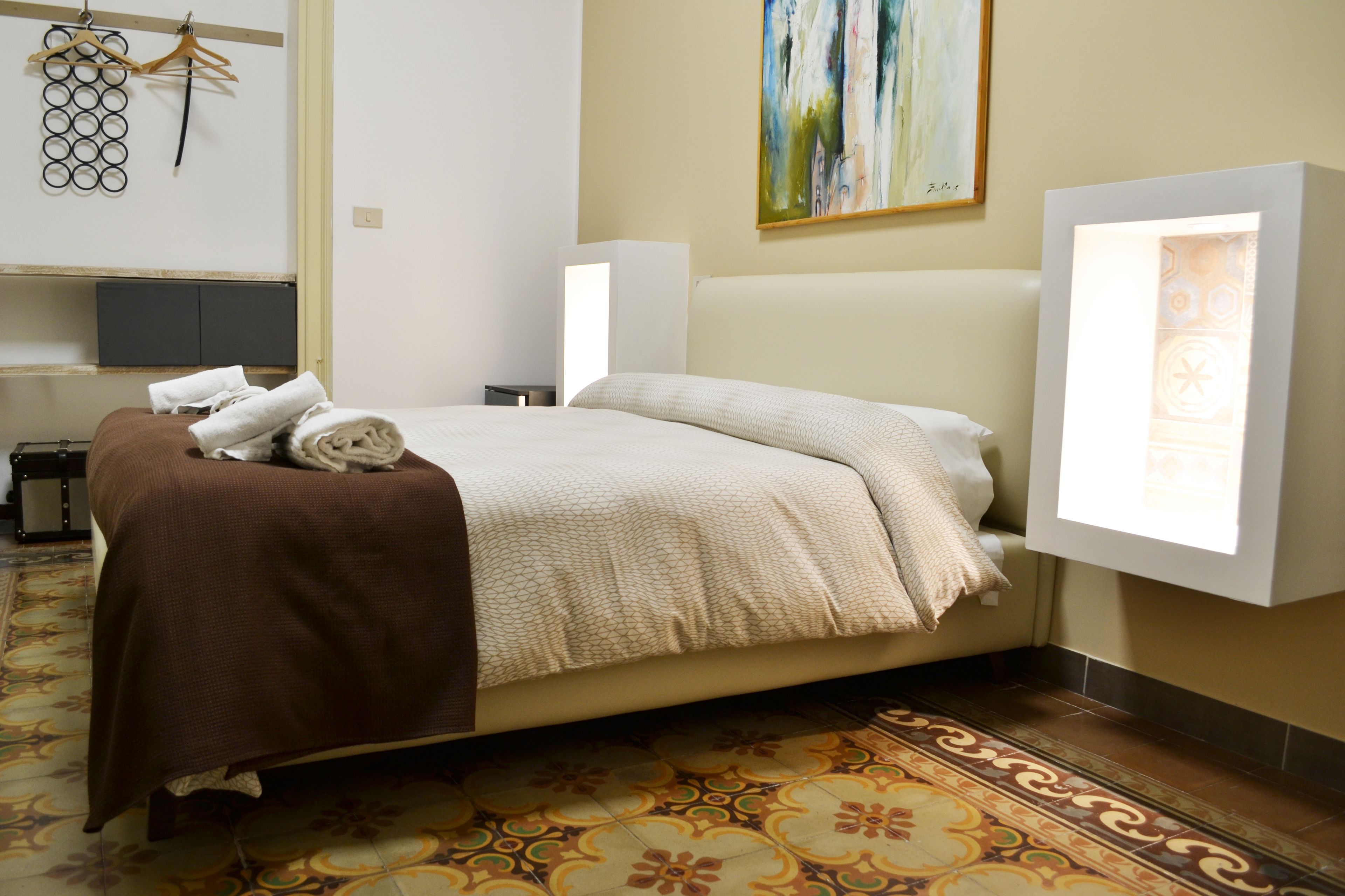 Ein ordentlich bezogenes Bett mit einer braunen Decke, einem beigen Bettüberwurf und einem weißen Kopfkissen. Darüber hängt ein Bild an der Wand und zur Rechten befindet sich eine Wandleuchte.