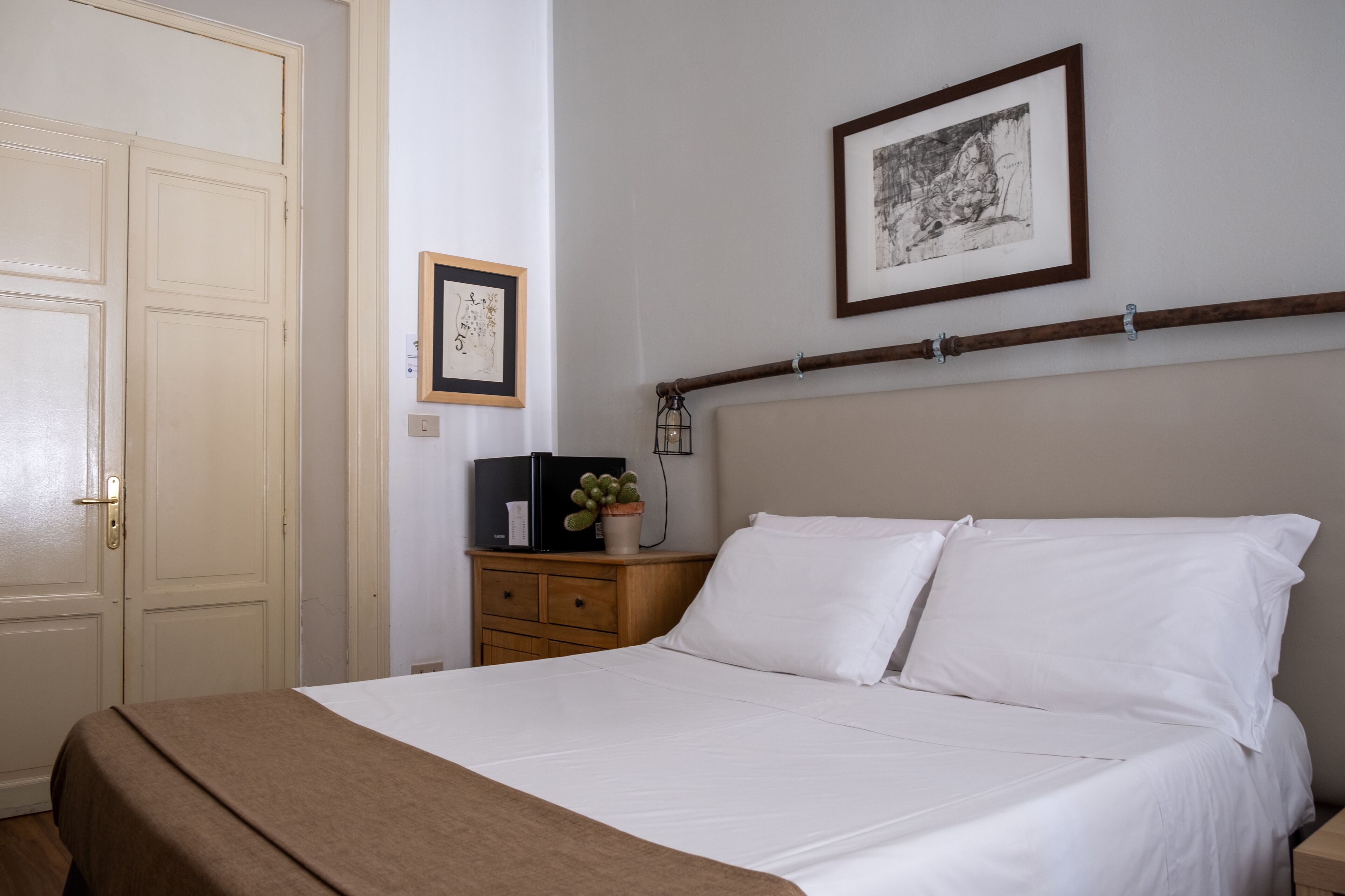 Una cama bien hecha con sábanas blancas, una mesita de noche con lámpara y un cuadro enmarcado en la pared.