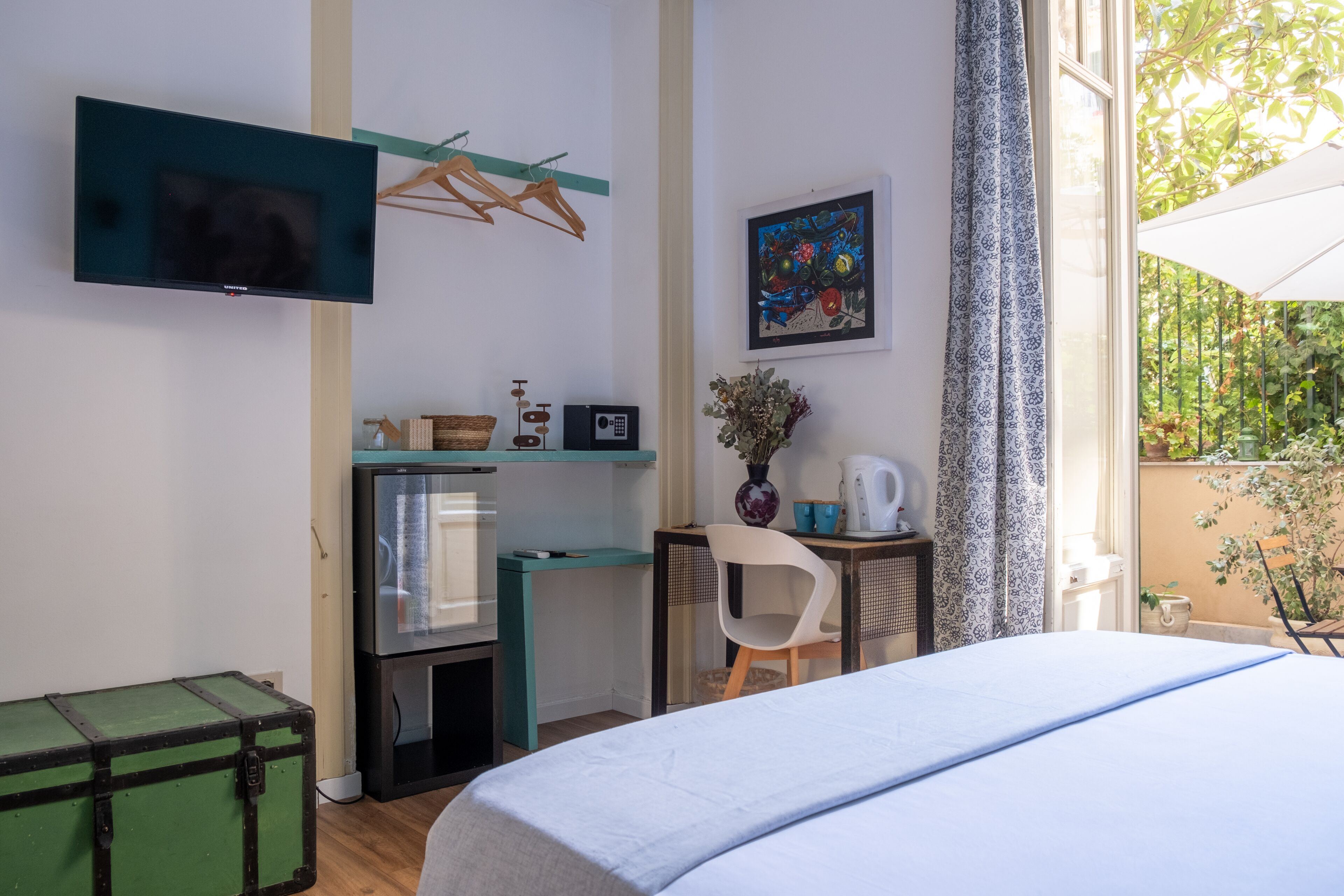 A bedroom with a bed, a desk, a TV, and a window with a view of greenery.