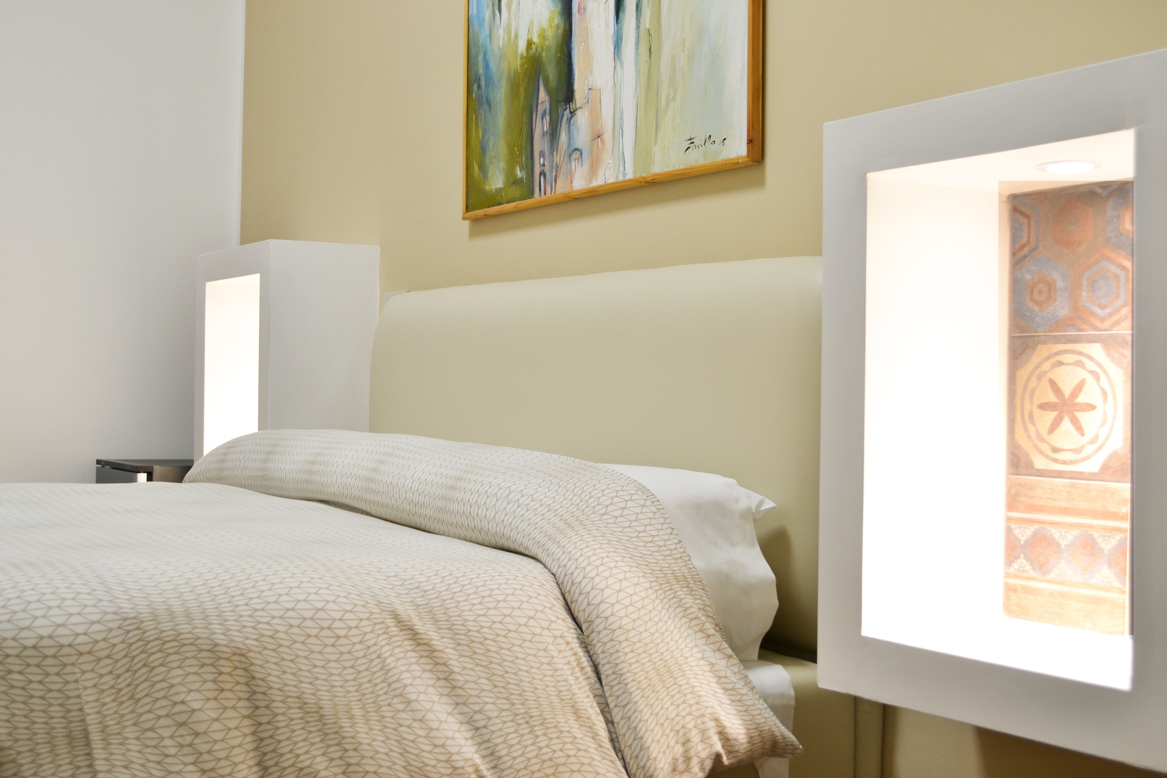 Ein Hotelzimmer mit Bett, Nachttisch, einem Bild an der Wand und einem Fenster mit gemusterten Vorhängen.