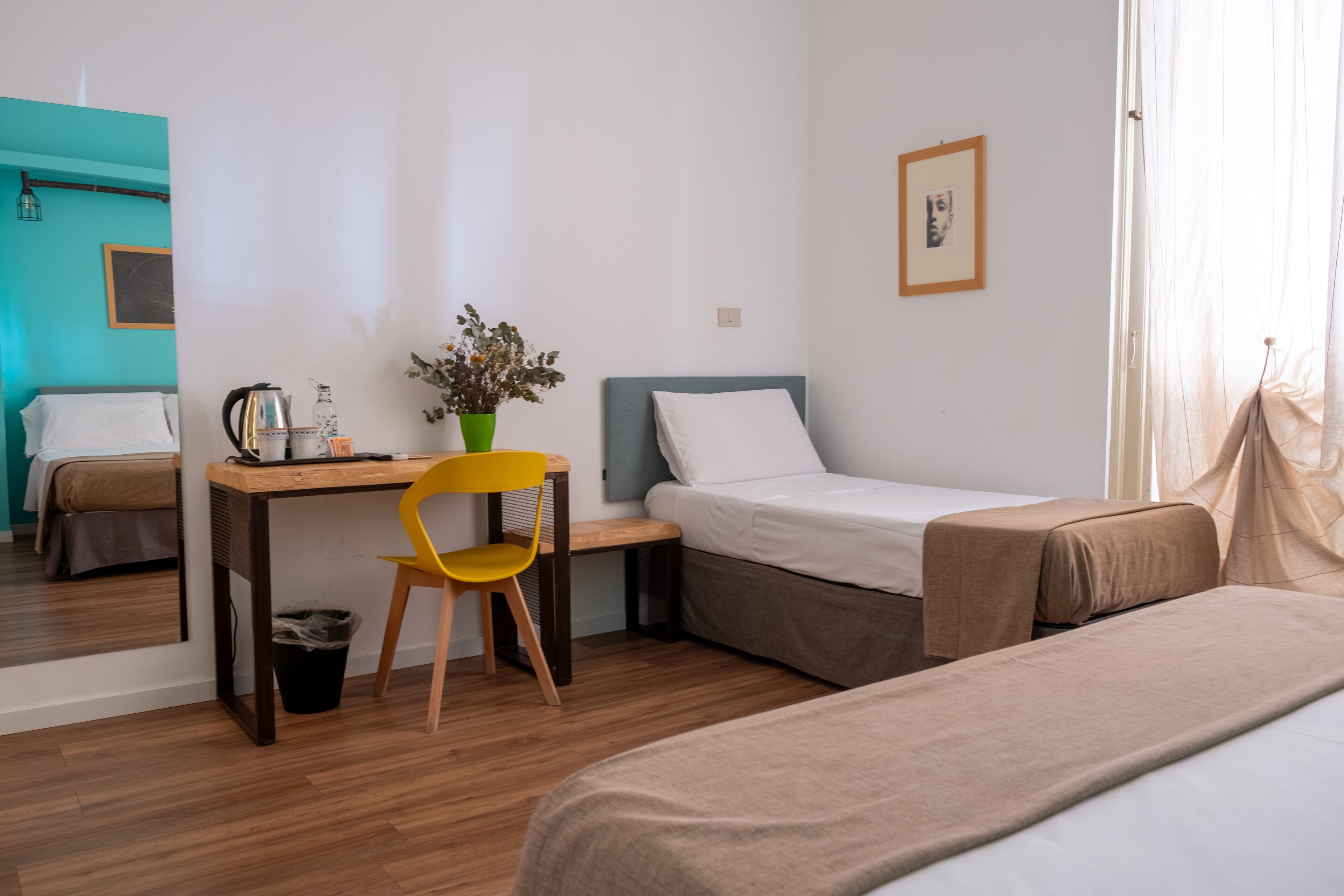 A hotel room with a bed, a desk with a chair, a small table with a plant, and a window with sheer curtains.