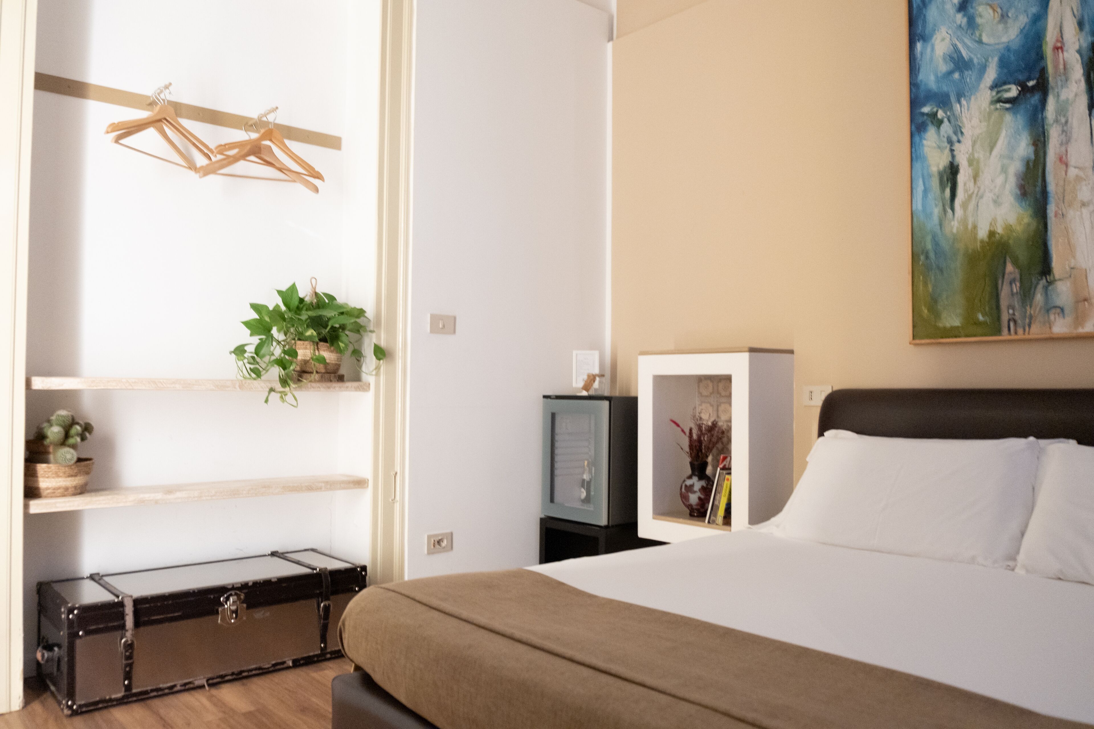 A bedroom with a bed, a wardrobe, a plant, and a painting on the wall.