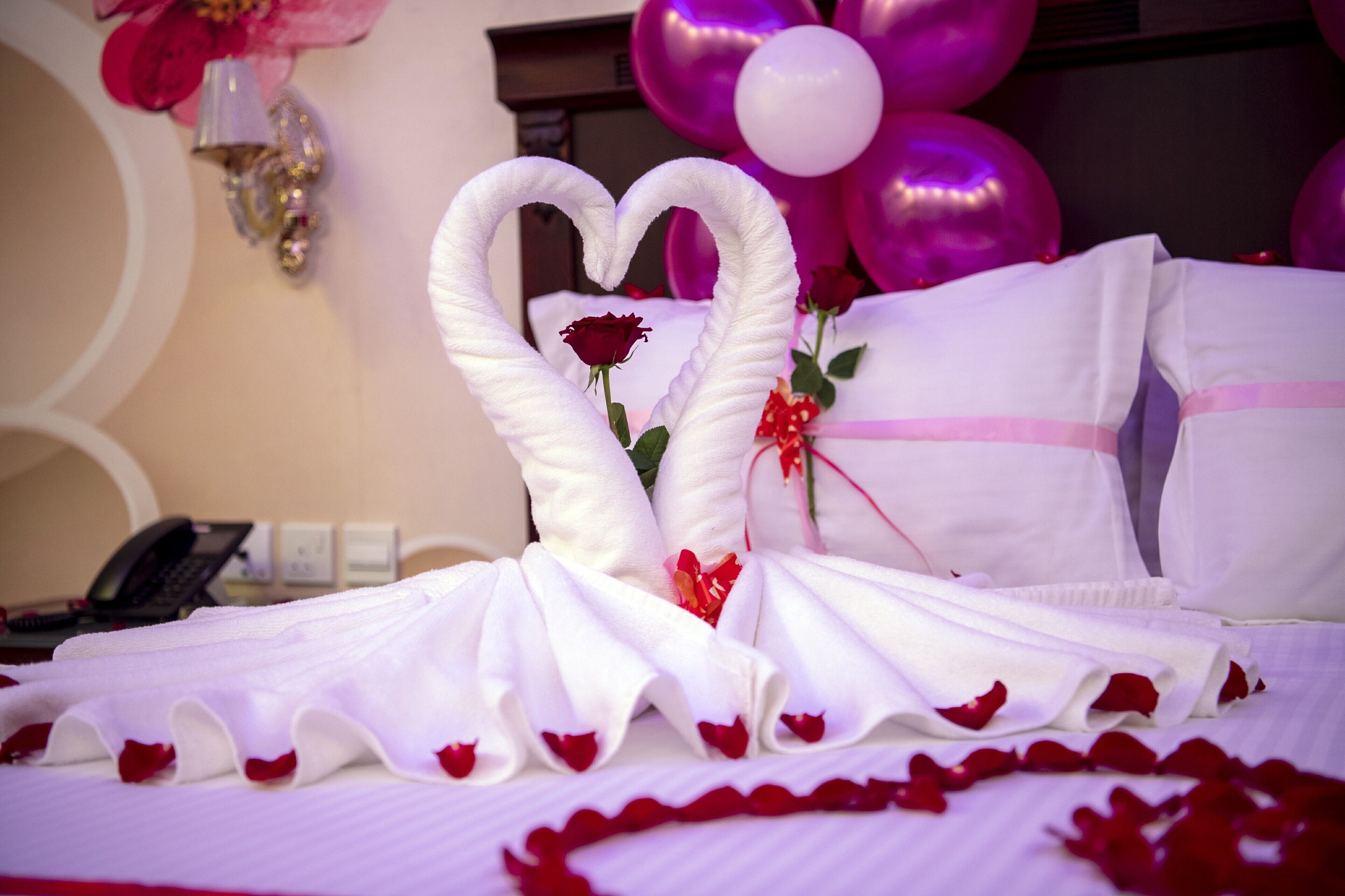 Una cama con sábanas blancas, una disposición de toallas en forma de corazón y pétalos de rosa.