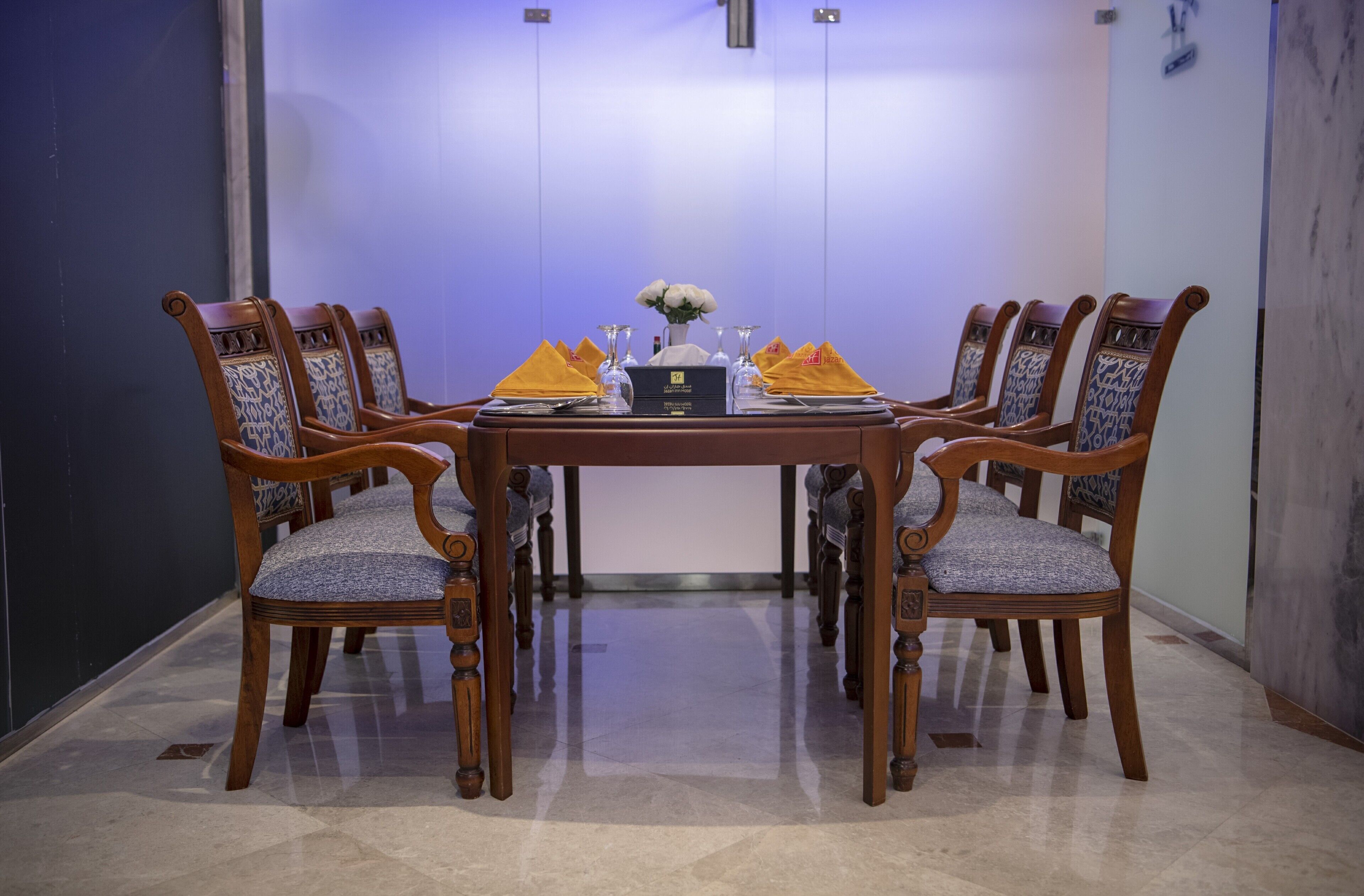 A conference room with a round table, six wooden chairs with patterned upholstery, and a vase with white flowers.