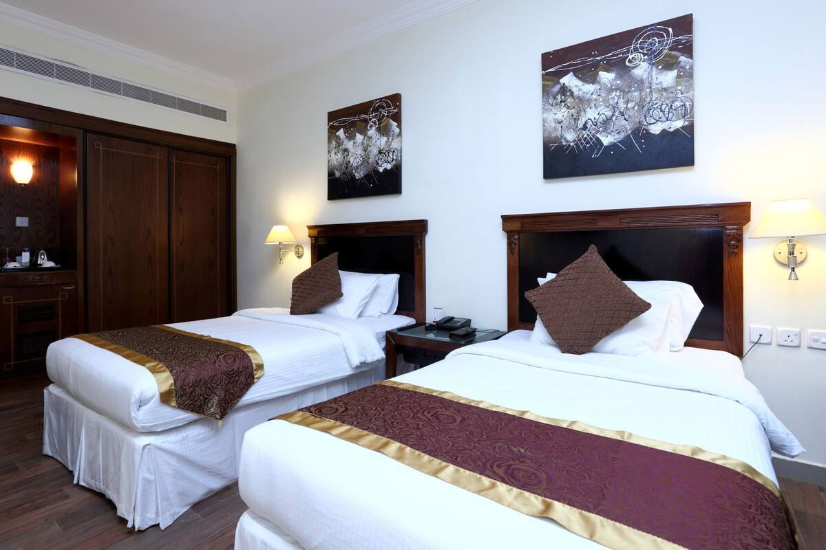 A hotel room with two beds, wooden headboards, a nightstand, a desk, a lamp, and artwork on the walls.