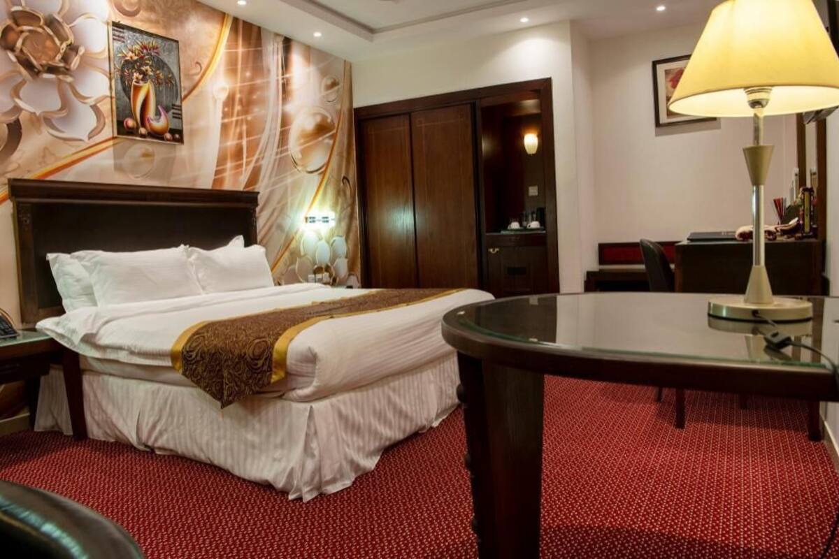 A hotel room with a bed, a desk, a lamp, a wardrobe, and a wall with a decorative painting.