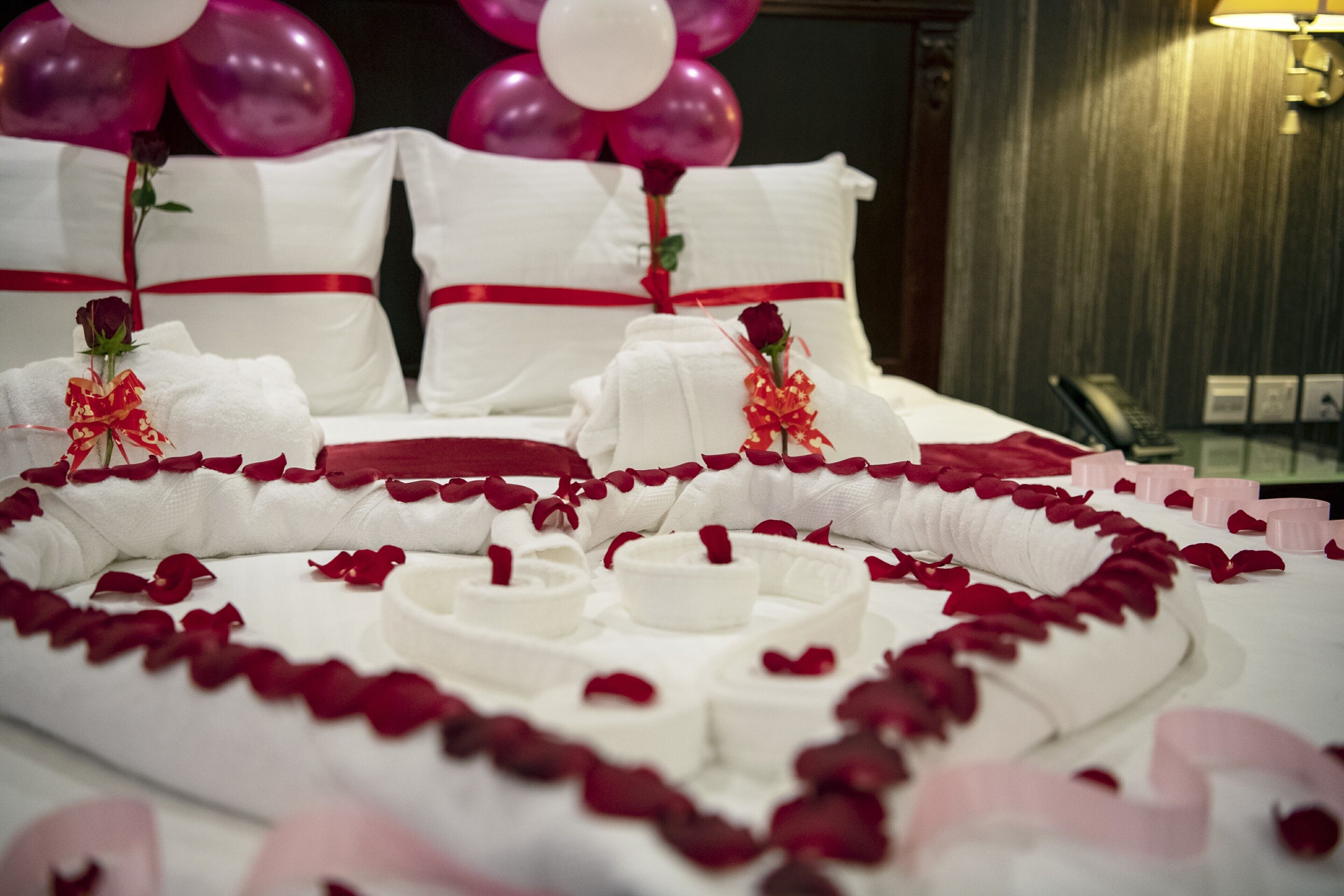 A bed with white linens, rose petals, and a heart-shaped arrangement of white candles.