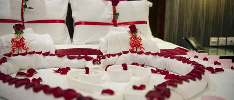 A bed with white linens, rose petals, and a heart-shaped arrangement of white candles.