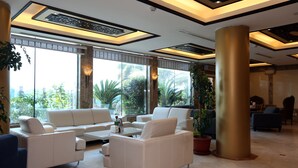 A spacious lobby with a modern design, featuring a large seating area with white sofas and a coffee table, large windows offering a view of palm trees, and a decorative ceiling with geometric patterns. - Jazan Inn (Jizan)