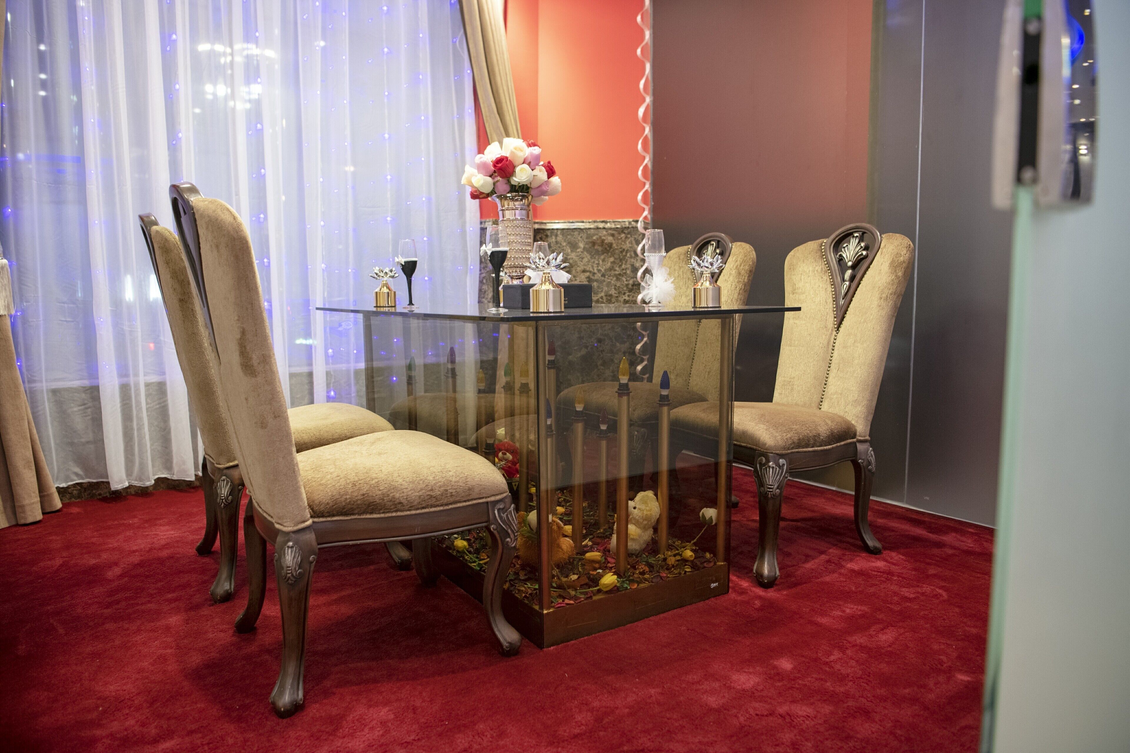Uma mesa de jantar com tampo de vidro, acompanhada de quatro cadeiras estofadas, um vaso com flores e um aparador de vidro decorado, com um urso de pelúcia e outros objetos.