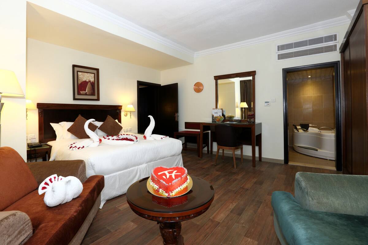 A hotel room with a bed, a desk, a chair, a table with a cake, and a bathroom.
