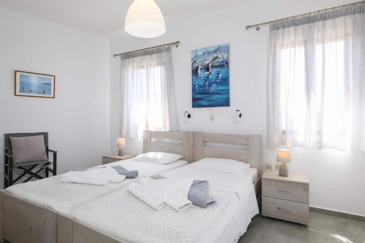 A double bed with white linens, two bedside tables with lamps, a chair, and a painting of boats on the wall.