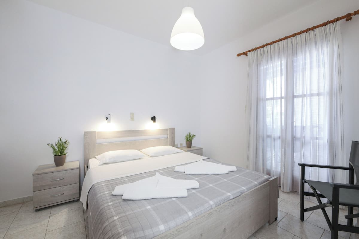 A modern bedroom with a large bed, bedside tables with plants, a desk, and a window with curtains.
