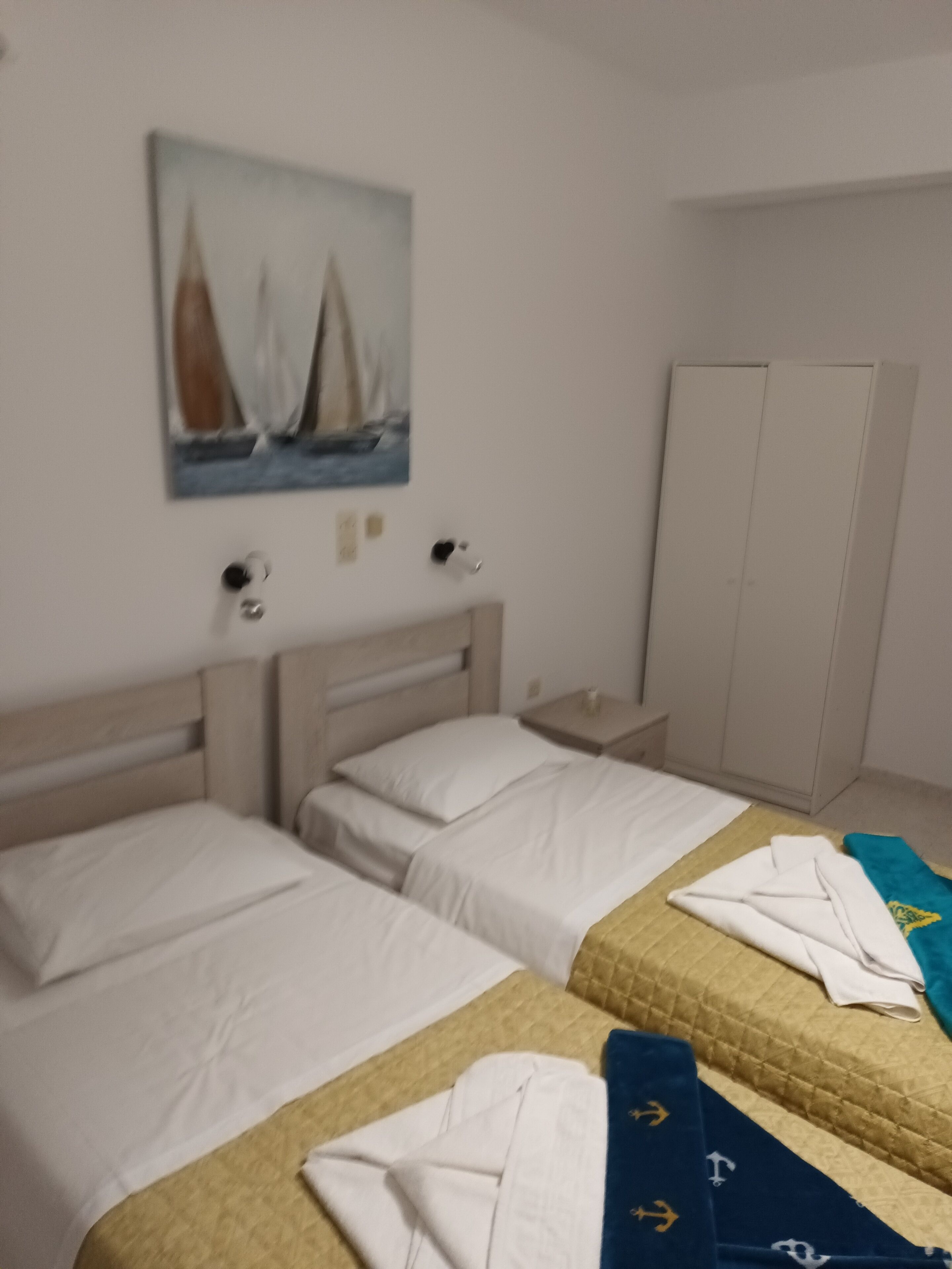 Quarto de hotel com duas camas, um guarda-roupa, um quadro na parede e uma mesinha com abajur.