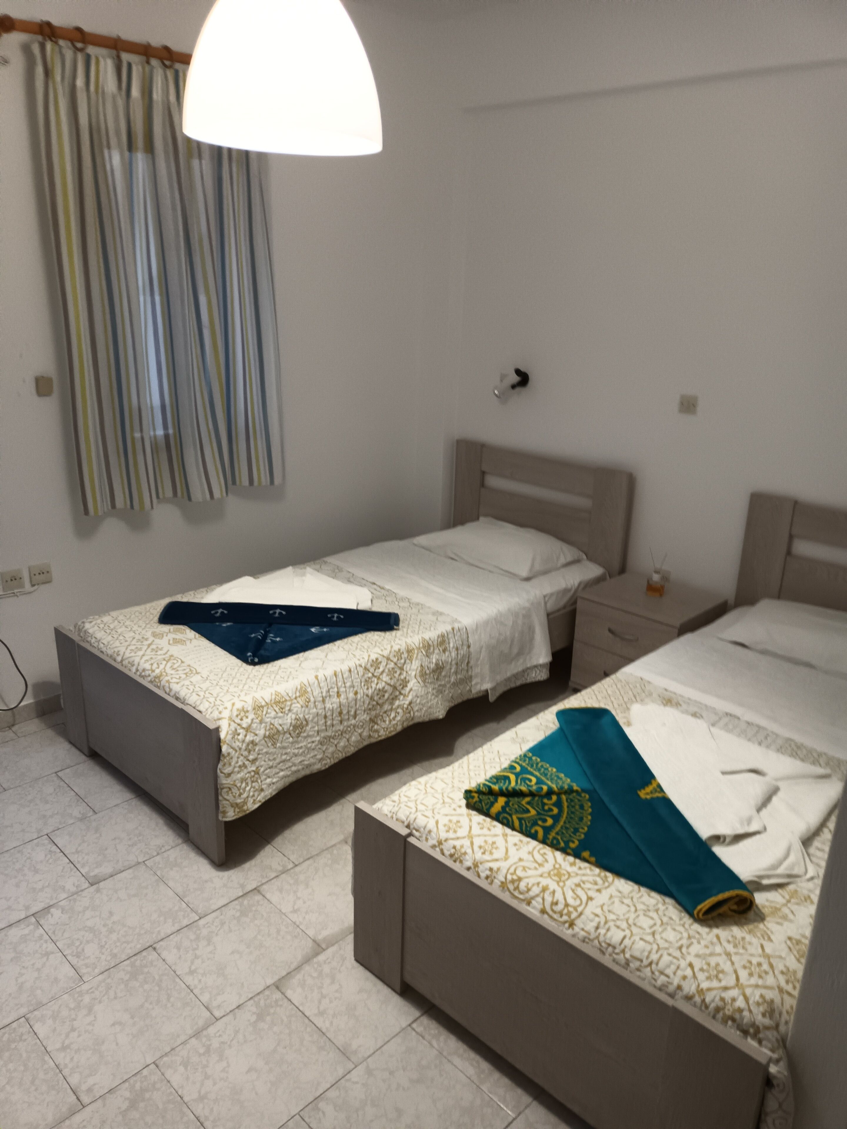 Un dormitorio con dos camas individuales, una mesita de noche con lámpara, y una ventana con cortinas.