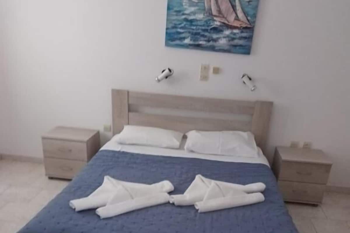 A neatly made bed with a blue bedspread and white pillows, two bedside tables, and a painting of sailboats on the wall.