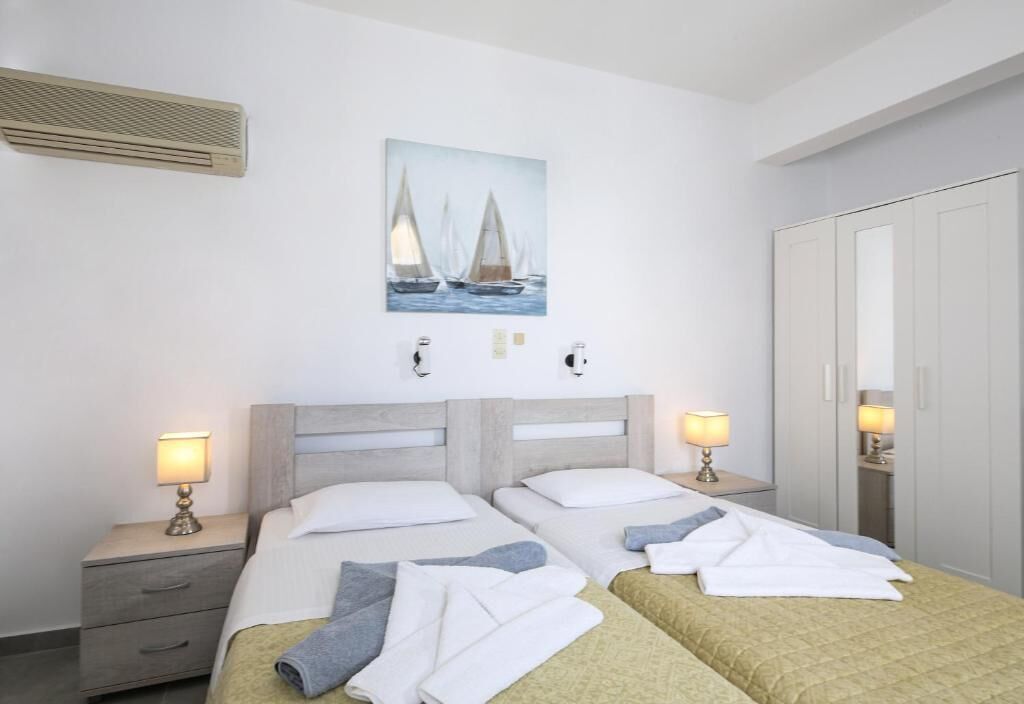A hotel room with two beds, a painting of sailboats, bedside lamps, a wardrobe, and a heater.