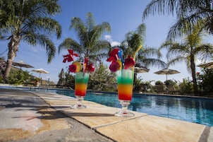 Dos cócteles con adornos de frutas sobre una mesa junto a la piscina, rodeados de palmeras y sombrillas.