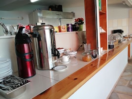 Montaje de un bufé de desayuno con una cafetera de acero inoxidable, una botella de agua roja y diversos alimentos sobre una encimera de madera.