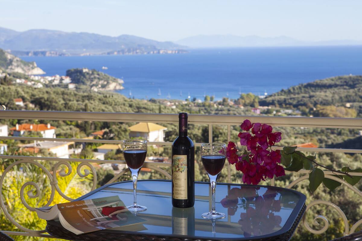 A table with a bottle of wine and two glasses on a balcony overlooking a coastal landscape.