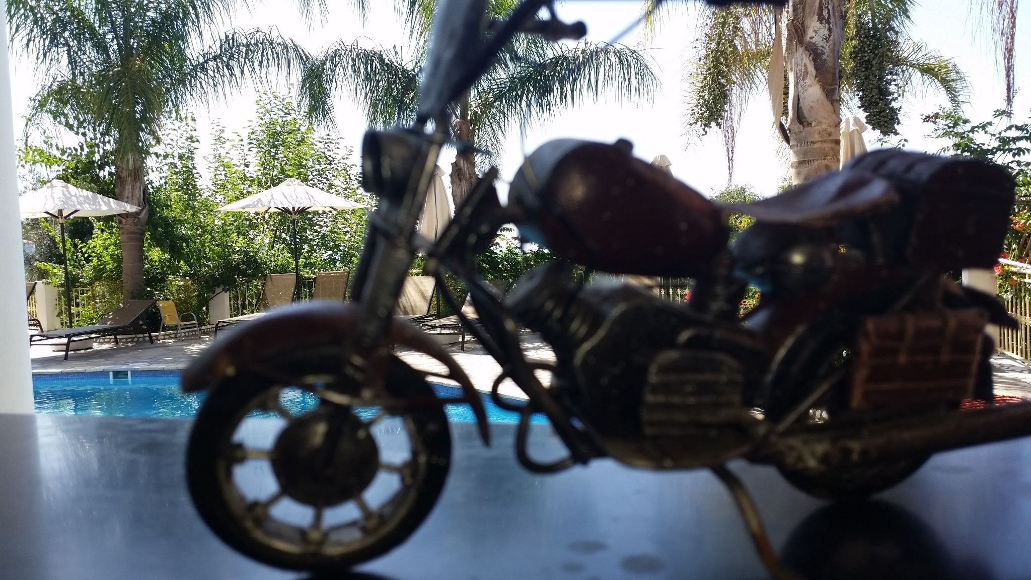 Una motocicleta modelo en un área junto a la piscina, con vista despejada a la piscina, sillas de descanso y palmeras.