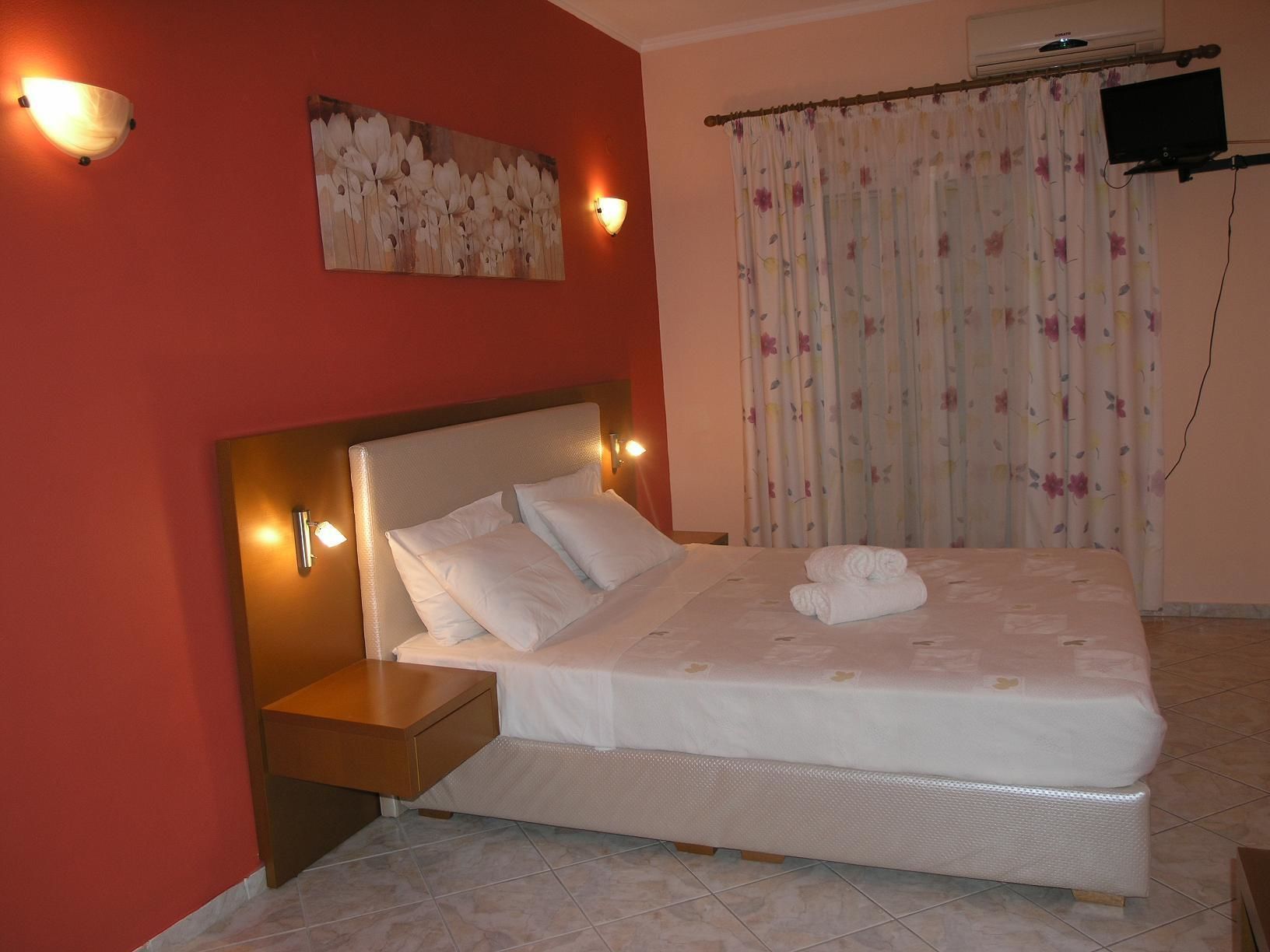 Una cama bien hecha con sábanas blancas y una toalla doblada, una mesita de noche de madera con una lámpara, un cuadro en la pared y una ventana con cortinas florales.