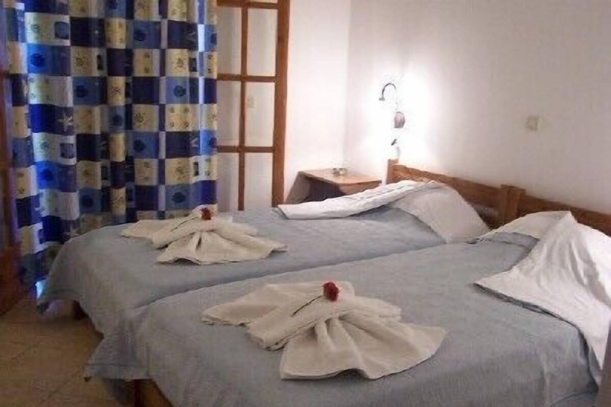 A room with two beds, each with a folded white towel and a red flower. There is a window with a checkered curtain, a nightstand with a lamp, and a wooden headboard.