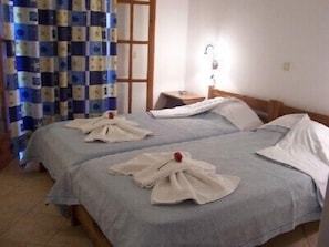 Una habitación con dos camas, cada una con una toalla blanca doblada y una flor roja. Hay una ventana con cortina a cuadros, una mesita de noche con una lámpara y un cabecero de madera.