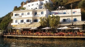 Ein weißes Gebäude mit Balkonen, einem Restaurantbereich mit Sonnenschirmen und Blick auf das Wasser.