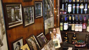 Ein Klavier mit gerahmten Fotografien davor, eine Weinbottelsammlung und eine Wand aus Holzpaneelen.