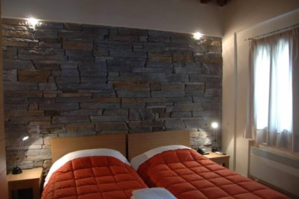 Een slaapkamer met een stenen muur, twee bedden met oranje beddengoed, bedlampen en een raam met gordijnen.