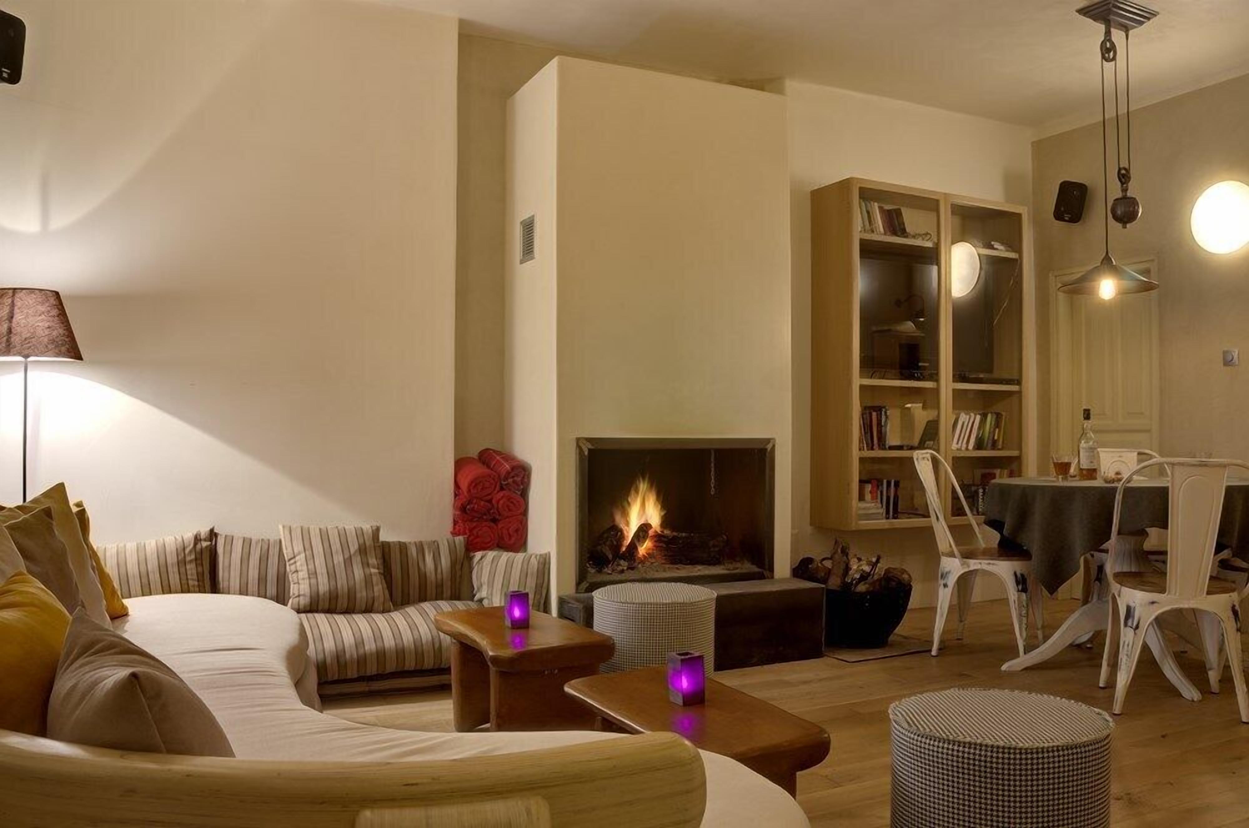 A cozy living room with a fireplace, a sofa, a dining table with chairs, and a bookshelf.