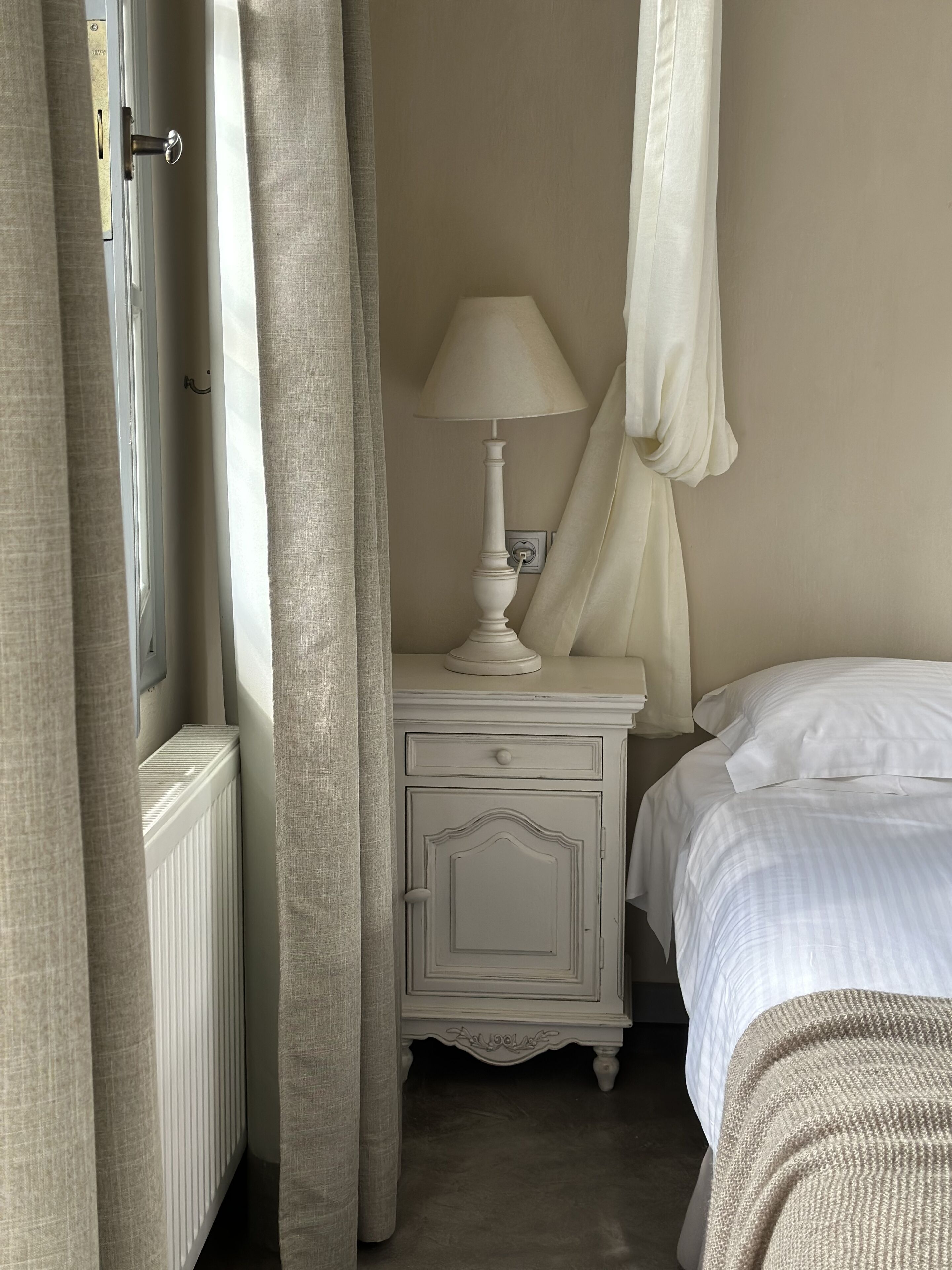 A bedroom with a bed, a nightstand with a lamp, a radiator, and curtains.