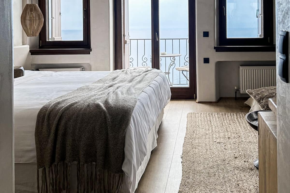 Ein Schlafzimmer mit einem großen Bett, Meerblick durch eine Schiebetür und ein Balkon mit einem Sessel.