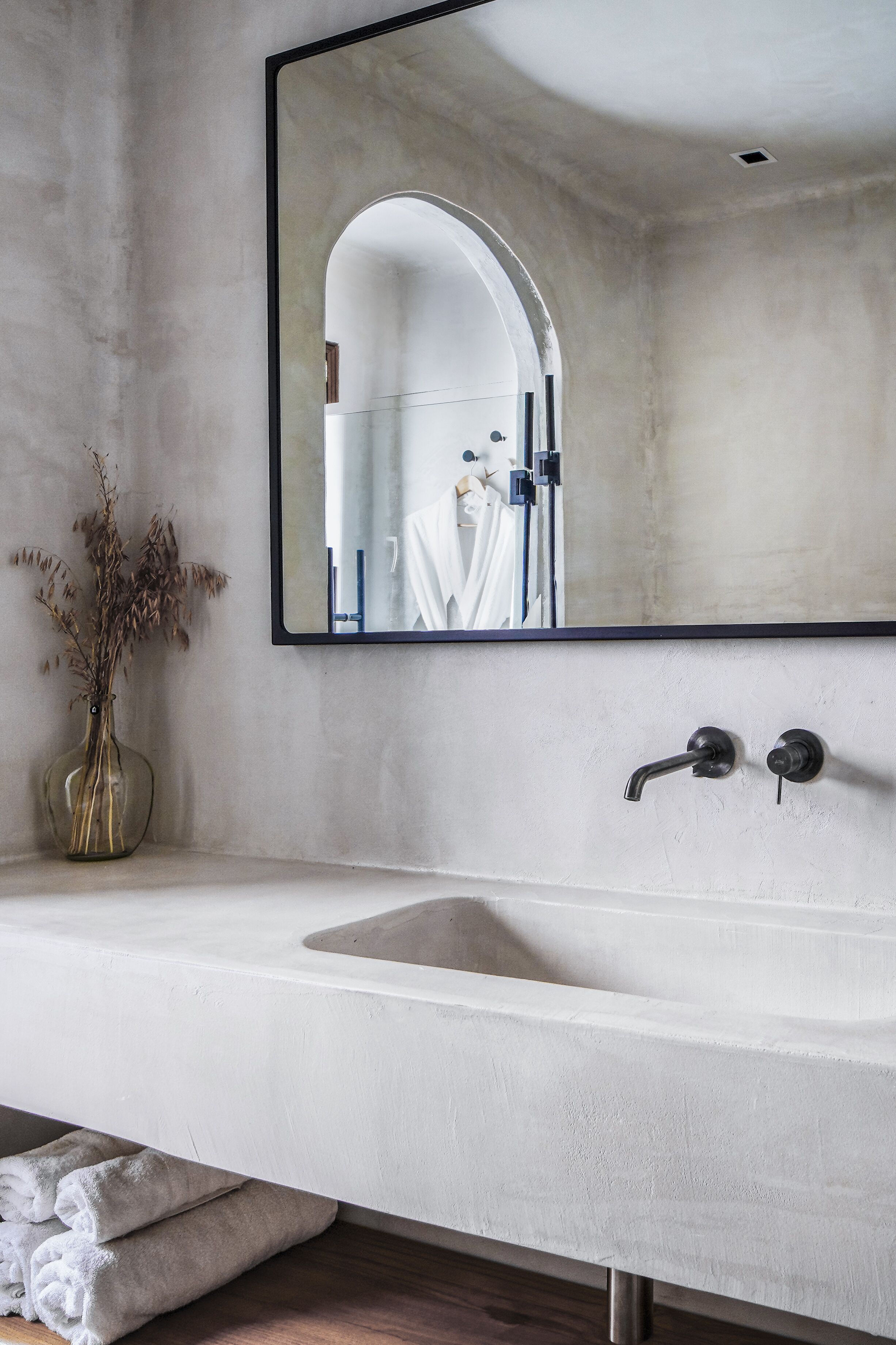 Un baño con un espejo grande, un lavabo blanco y un florero con flores secas.