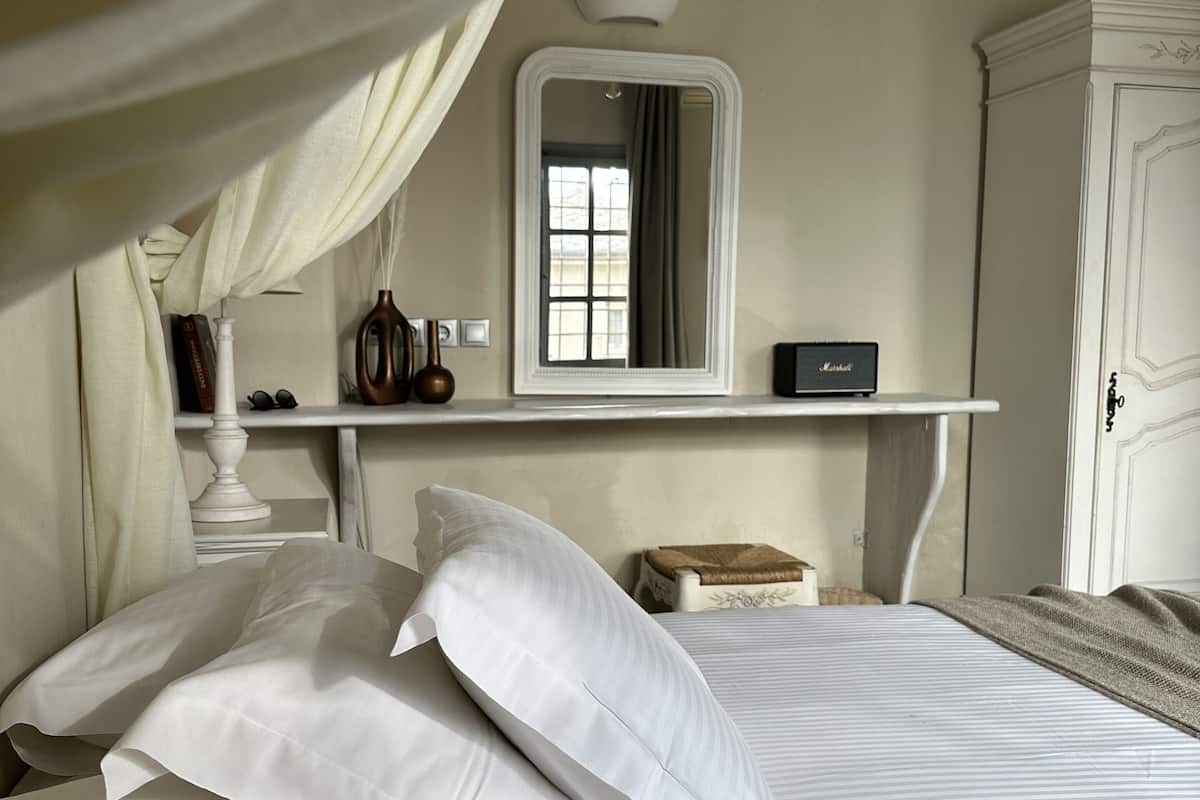 Ein ordentlich bezogenes Bett mit weißen Leinen und Kissen, ein Nachttisch mit Lampe, Spiegel und einem kleinen Regal mit Büchern und einer Vase.
