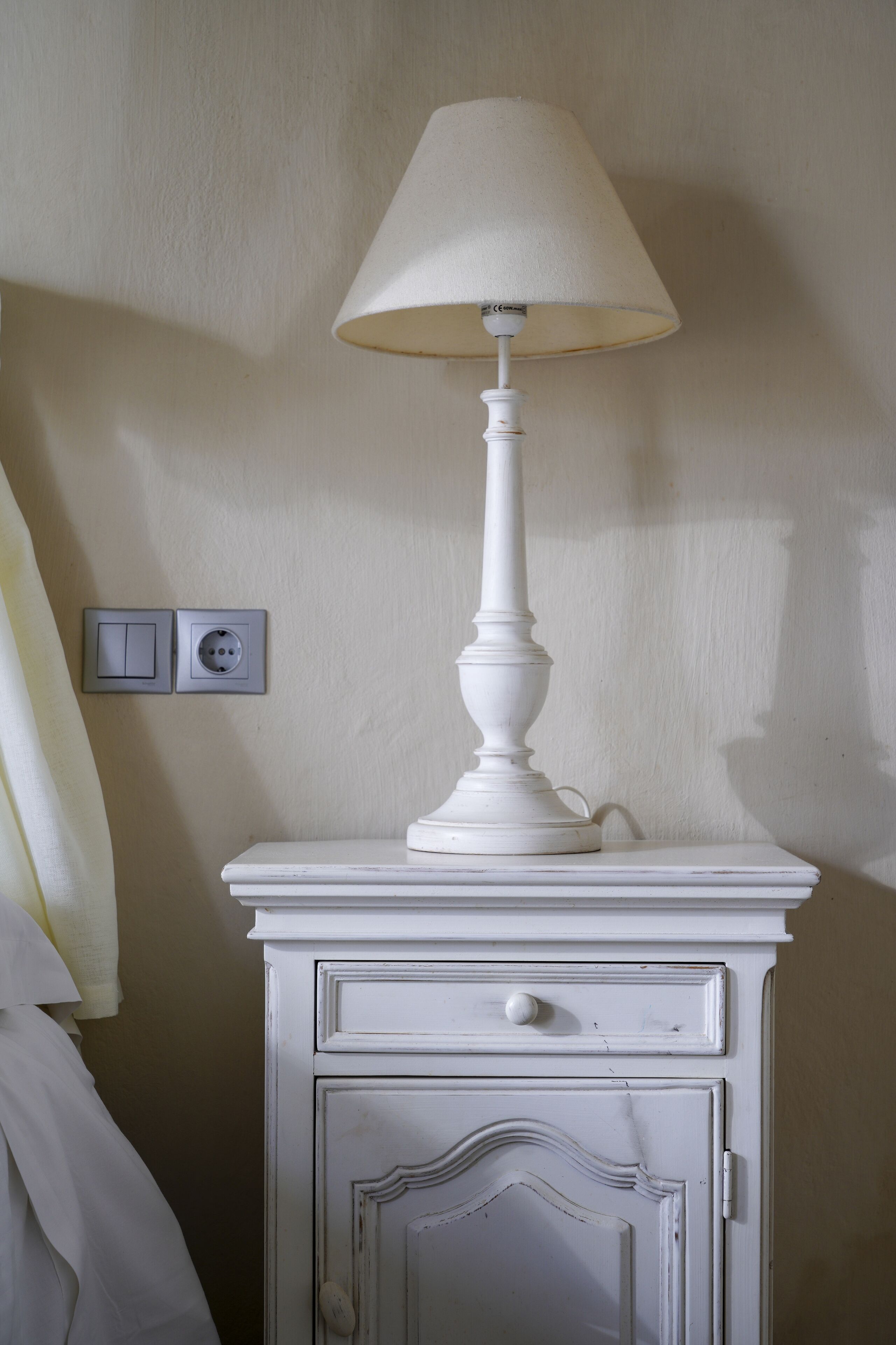 A white bedside table with a lamp, a drawer, and a switchboard with two switches and a socket.